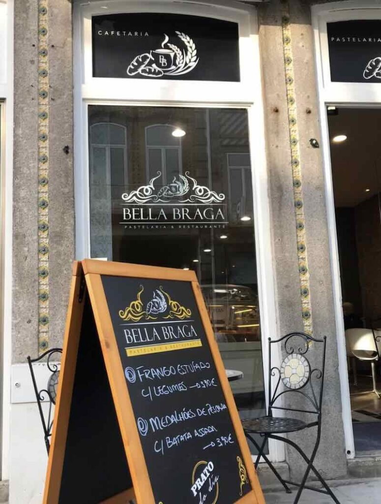 Exterior photo of Bella Braga with logo on window and black cafe table outside. Chalkboard aframe sign displays specials.